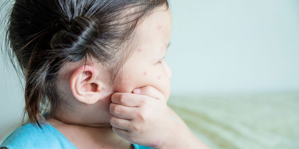 Ein Kind mit dunklen Haaren, das zu einem Zopf gebunden ist, trägt ein hellblaues Oberteil. Im Hintergrund ist eine weiße Wand und ein unscharfer Bereich in grünlichen Tönen zu sehen.