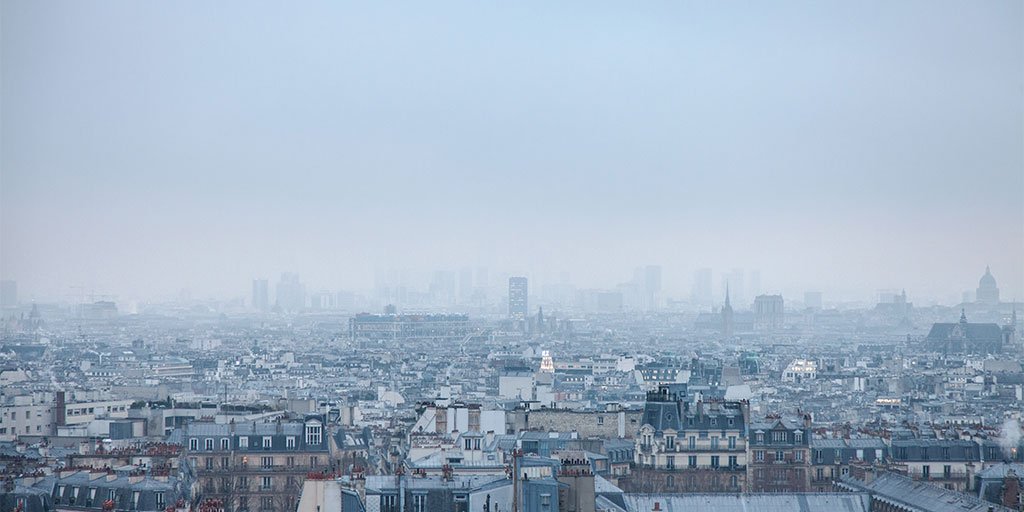 Blick über die Dächer von Paris an einem trüben, nebligen Tag. Im Vordergrund sind zahlreiche Häuser und Gebäude im typischen Pariser Stil mit Schieferdächern und Schornsteinen zu sehen. Im Hintergrund verschwimmen die Konturen moderner Wolkenkratzer und historischer Gebäude durch den dichten Nebel.