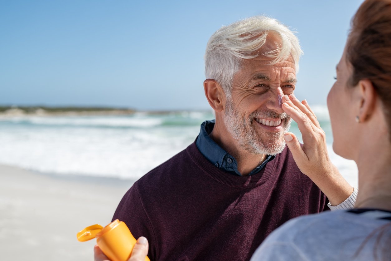 Frau cremt die Nase eines Mannes mit grauen Haaren und Falten im Gesicht am Strand ein