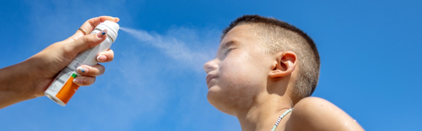 Hand sprüht Sonnenschutzspray auf das Gesicht eines Kindes vor blauem Himmel 