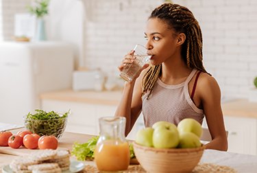 Junge Frau trinkt Wasser und vor ihr steht Gemüse und Obst