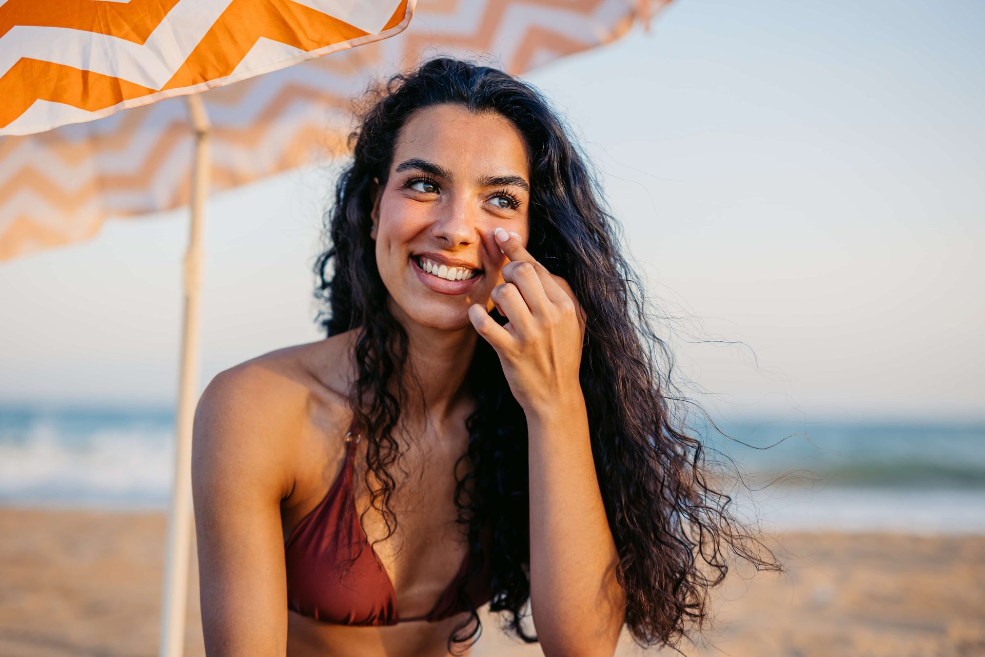 Lächelnde Frau trägt am Strand unter einem Sonnenschirm Creme im Gesicht auf