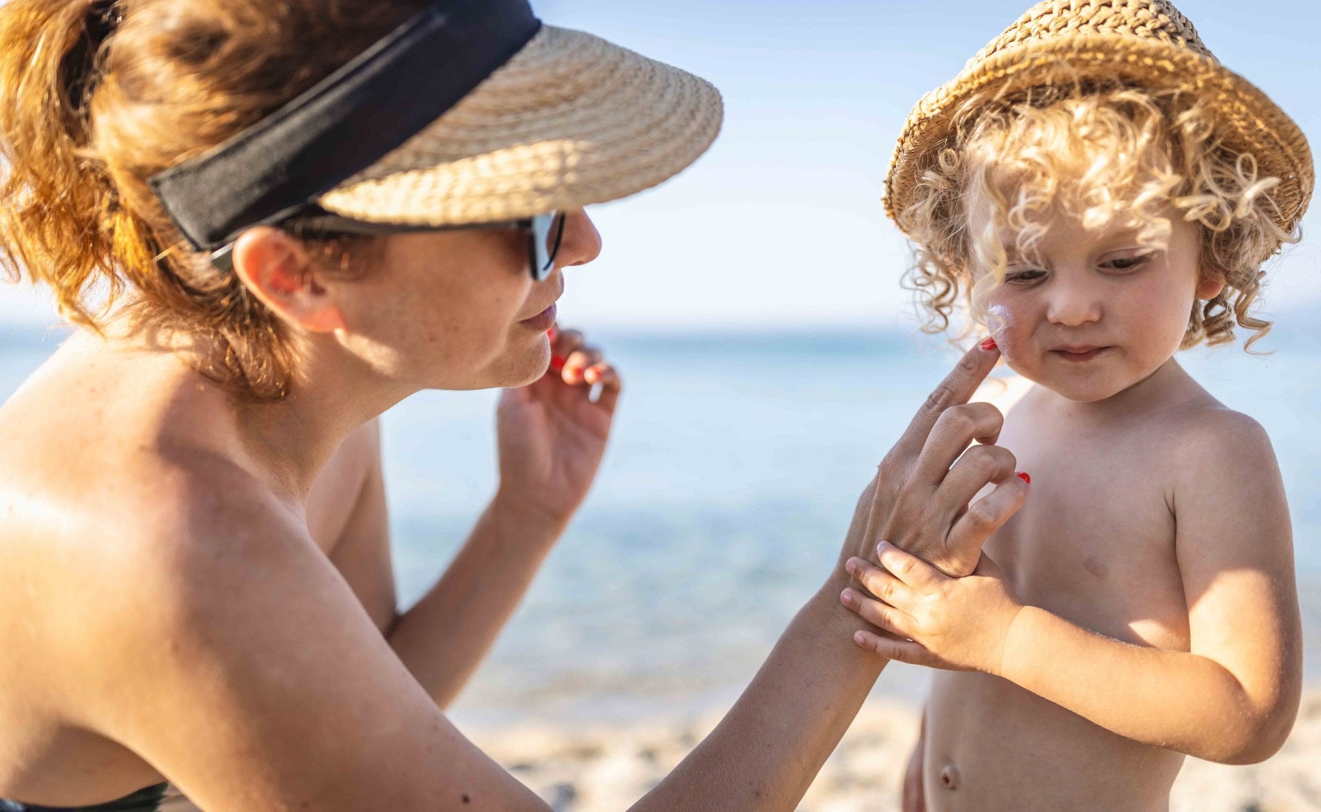 Mutter trägt einem kleinen Kind am Strand Sonnencreme auf die Wange auf