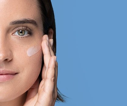 Leicht lächelnde Frau vor blauem Hintergrund trägt Creme unter ihrem Auge auf 