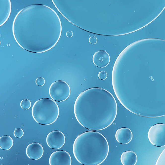 Schimmernde, transparente Flüssigkeitstropfen auf hellem Hintergrund, die den Inhaltsstoff ultrafeines Öl darstellen und für eine leichte, nährende Textur stehen.