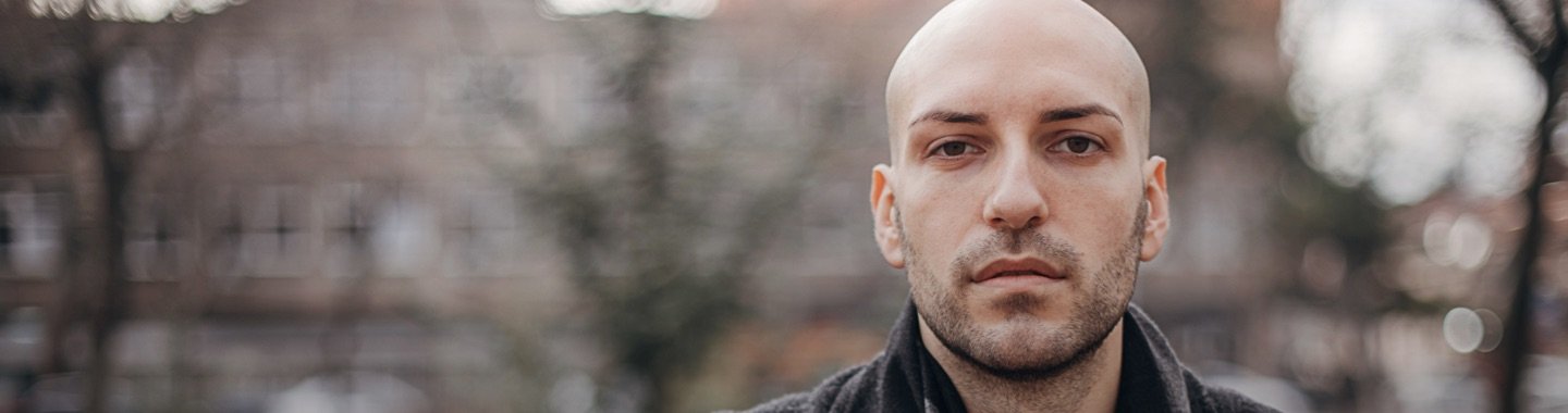 Eine Person steht draußen vor einem unscharfen Hintergrund mit Bäumen und Gebäude. Winterliche oder herbstliche Umgebung.